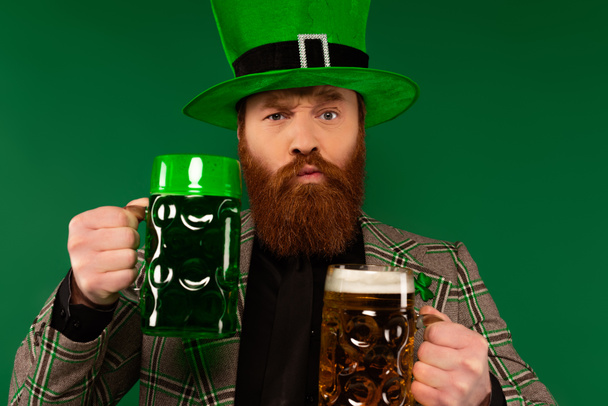 Hombre reflexivo en sombrero sosteniendo vasos de cerveza y mirando a la cámara aislada en verde  - Foto, imagen