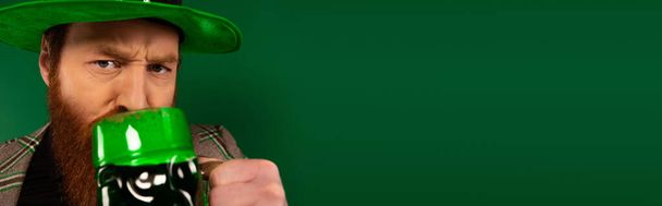 Σοβαρός άνδρας με καπέλο πίνοντας μπύρα ενώ γιορτάζει την ημέρα του Αγίου Πατρικίου απομονωμένος στο πράσινο, πανό   - Φωτογραφία, εικόνα