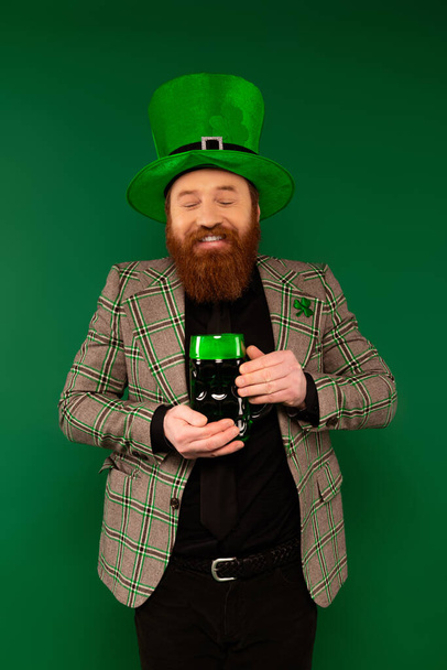 Joyful bearded man holding glass of beer while celebrating saint patrick day isolated on green  - 写真・画像