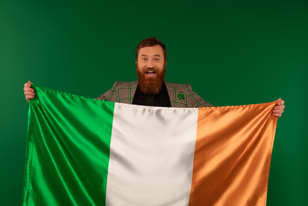 Feliz hombre barbudo sosteniendo bandera irlandesa y mirando a la cámara aislada en verde  - Foto, Imagen