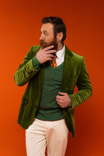 Модный человек в бархатной куртке трогает бороду на красном фоне  - Фото, изображение