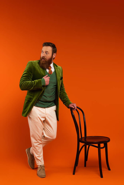 Full length of stylish man in jacket standing near chair on red background  - Foto, immagini