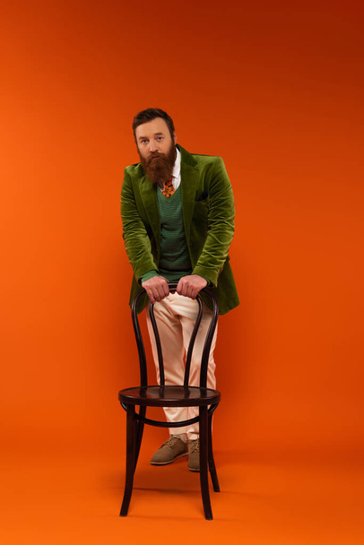Stylish bearded model looking at camera near chair on red background  - 写真・画像