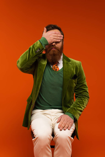 Trendy bearded man in jacket covering eyes while sitting on chair on red background  - Fotó, kép