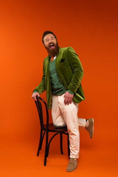 Cheerful bearded model in velvet jacket laughing near chair on red background  - Foto, afbeelding