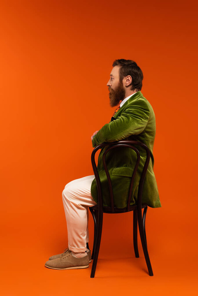 Side view of stylish man in velvet jacket sitting on wooden chair on red background  - Foto, afbeelding