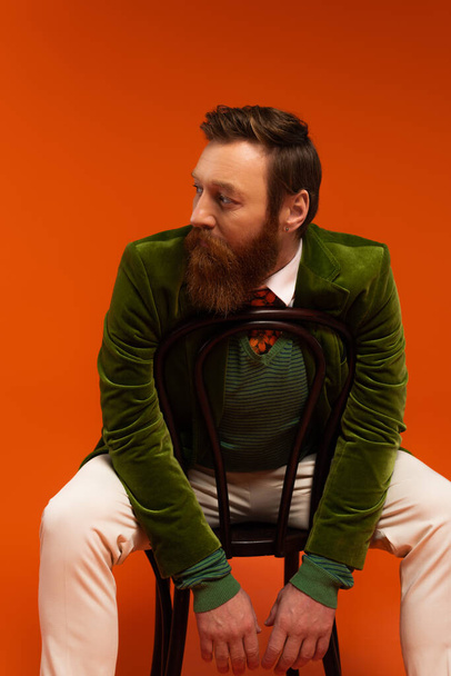 Trendy bearded man in green jacket sitting on chair on red background  - Photo, Image