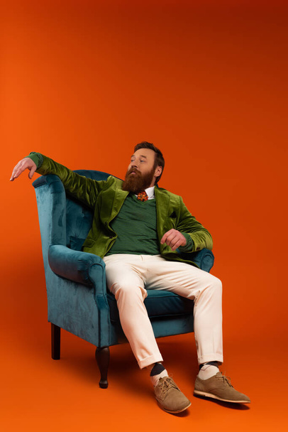 Стильный бородатый мужчина сидит на синем кресле на красном фоне  - Фото, изображение
