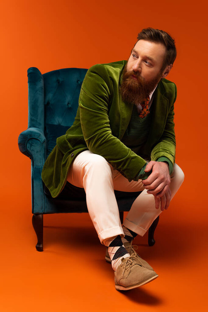 Trendy bearded man sitting on blue armchair on red background  - Foto, Imagen