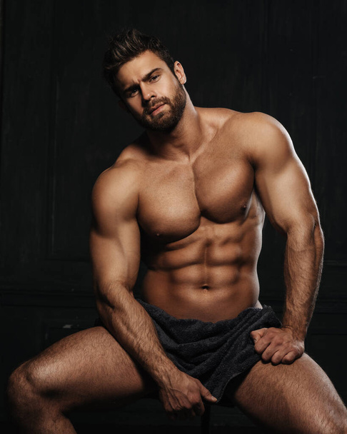 Brutal hombre sexy con seis abdominales paquete en toalla en el fondo negro. Guapo modelo de fitness masculino sentado en el estudio. barbudo musculoso chico cubierta con toalla en estudio. - Foto, imagen