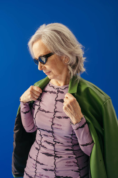 ブルーに隔離された緑の革のジャケットを着たスタイリッシュなサングラスの先輩女性  - 写真・画像