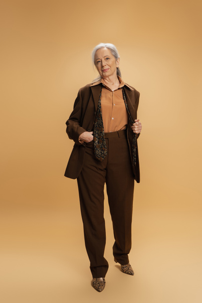 pleine longueur de la femme âgée en brun tenue formelle debout avec la main dans la poche sur fond beige  - Photo, image