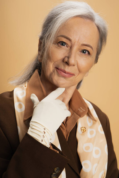 портрет довольной пожилой женщины в белой перчатке и коричневом костюме, смотрящей на камеру, изолированную на бежевом  - Фото, изображение
