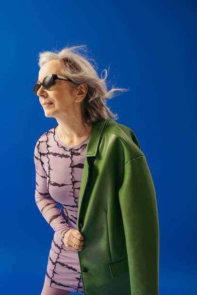 senior woman in dress and trendy sunglasses standing with green jacket isolated on blue  - Fotó, kép