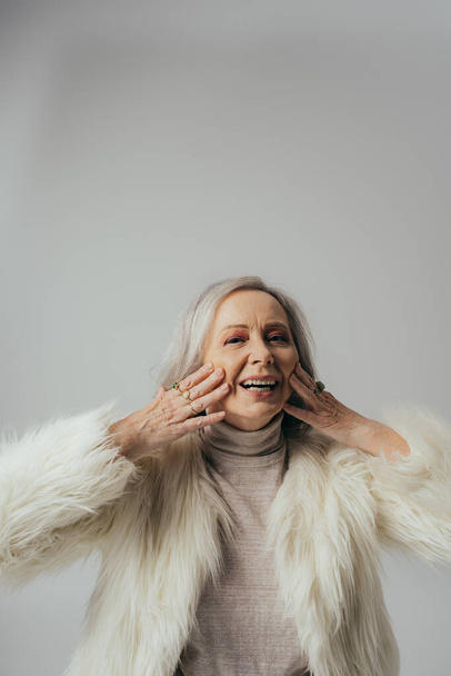 портрет веселой пожилой женщины с кольцами на пальцах, смотрящей в камеру при прикосновении лица к серому - Фото, изображение