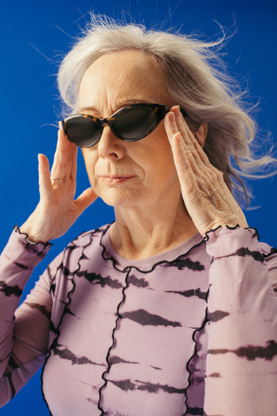 portrait of senior woman adjusting sunglasses isolated on blue  - Fotoğraf, Görsel