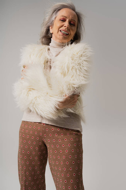 emotional senior woman in faux fur jacket posing with crossed arms isolated on grey  - Fotó, kép