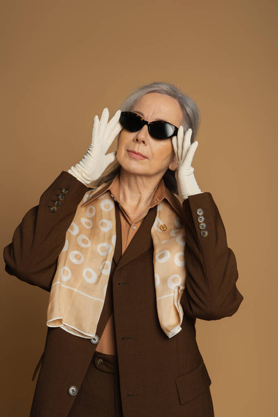 senior woman in brown formal wear and white gloves adjusting sunglasses isolated on beige  - 写真・画像