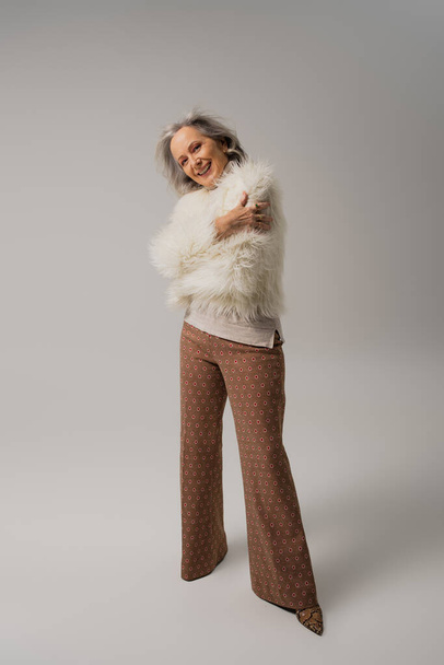 full length of happy elderly woman in white faux fur jacket posing on grey  - Фото, изображение