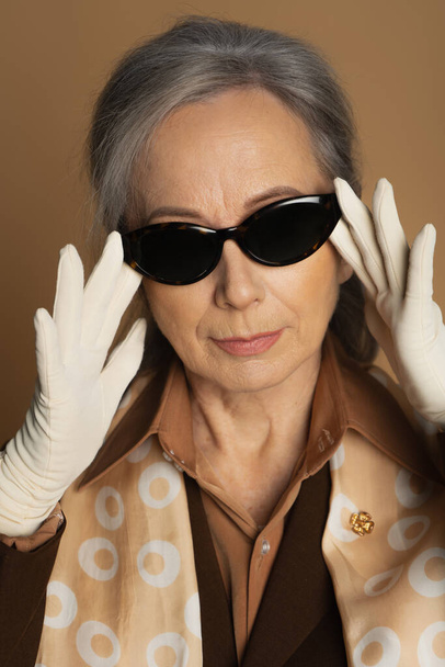 portrait of senior woman in white gloves adjusting sunglasses isolated on beige  - Foto, Imagen