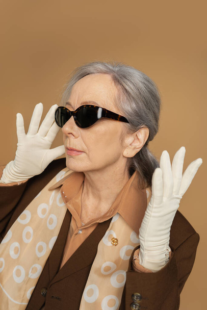 ηλικιωμένη γυναίκα με λευκά γάντια και γυαλιά ηλίου που ποζάρει απομονωμένη σε μπεζ  - Φωτογραφία, εικόνα