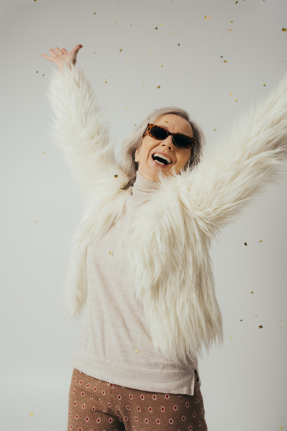 счастливая пожилая женщина в белом искусственном меховом пиджаке и солнцезащитных очках, поднимающих руки рядом с падающими конфетти на серый  - Фото, изображение