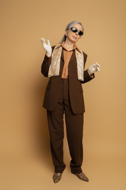 full length of senior woman in brown formal wear and white gloves standing on beige  - Fotografie, Obrázek