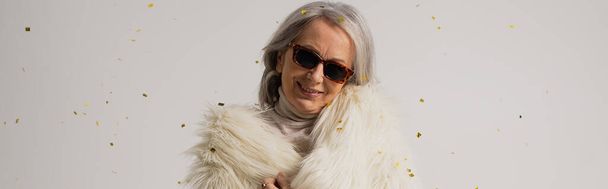 Beyaz sahte kürk ceketli ve güneş gözlüklü yaşlı bir kadın gri arka planda konfeti düşerken gülümsüyor.  - Fotoğraf, Görsel