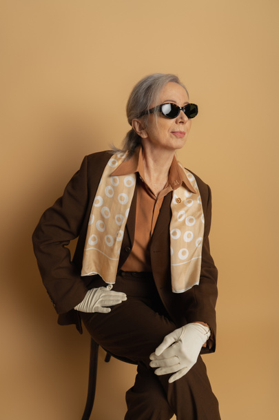 senior woman in brown suit and sunglasses sitting on wooden chair on beige  - Zdjęcie, obraz