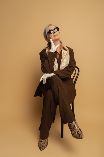 full length of thoughtful senior woman in brown formal wear and sunglasses sitting on wooden chair on beige  - Photo, image