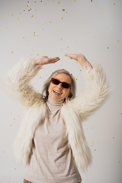 happy elderly woman in white faux fur jacket and sunglasses standing with raised hands near falling confetti on grey  - Foto, Imagen