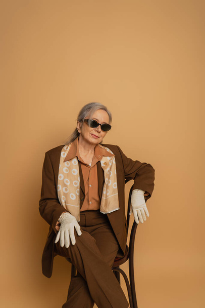 elderly woman in brown suit and sunglasses sitting on chair and looking at camera on beige  - Foto, Bild