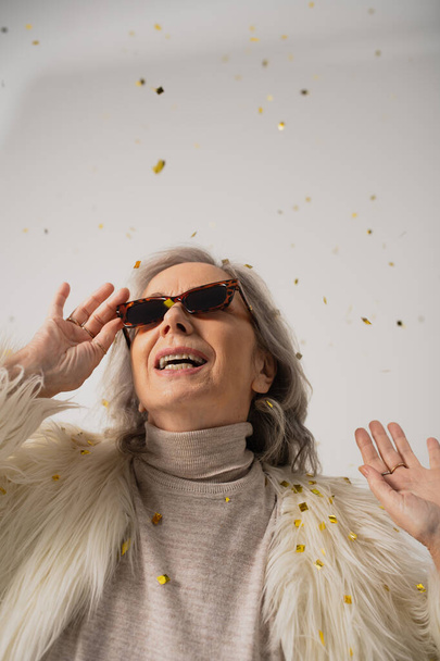 szczęśliwy starszy kobieta w biały faux futro kurtka i modne okulary słoneczne uśmiechając się blisko spadające konfetti na szarym tle  - Zdjęcie, obraz