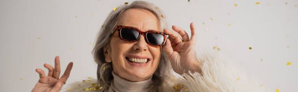 mujer mayor alegre ajustando gafas de sol de moda cerca de confeti caída sobre fondo gris, pancarta  - Foto, imagen