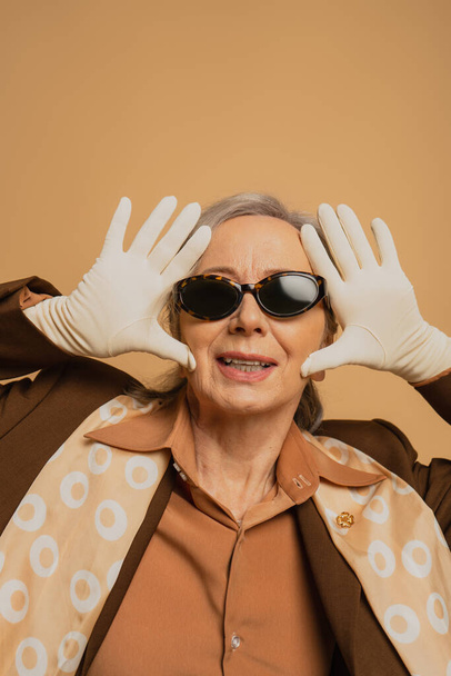 happy and stylish senior woman in sunglasses and white gloves posing isolated on beige  - Fotografie, Obrázek