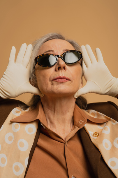 κομψή ηλικιωμένη γυναίκα με γυαλιά ηλίου και λευκά γάντια που ποζάρουν απομονωμένη σε μπεζ  - Φωτογραφία, εικόνα