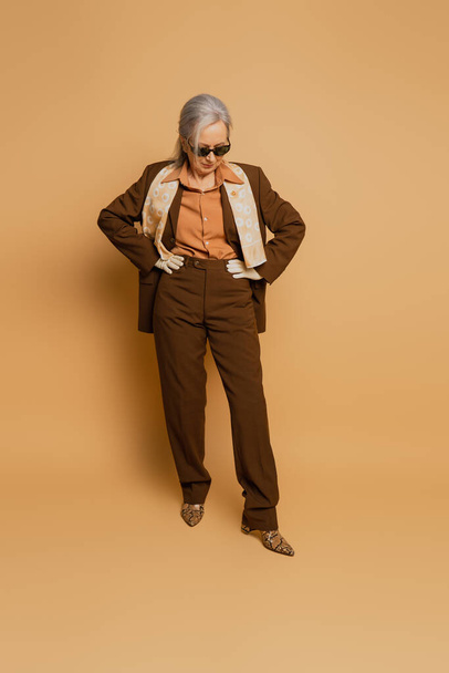 volle Länge der stilvollen Seniorin in Sonnenbrille und Anzug posiert mit den Händen auf den Hüften auf beige  - Foto, Bild