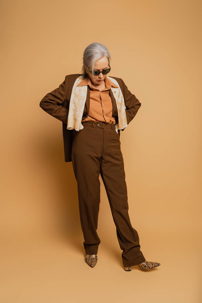 volledige lengte van stijlvolle oudere vrouw in zonnebril en pak poseren met de handen op heupen op beige  - Foto, afbeelding