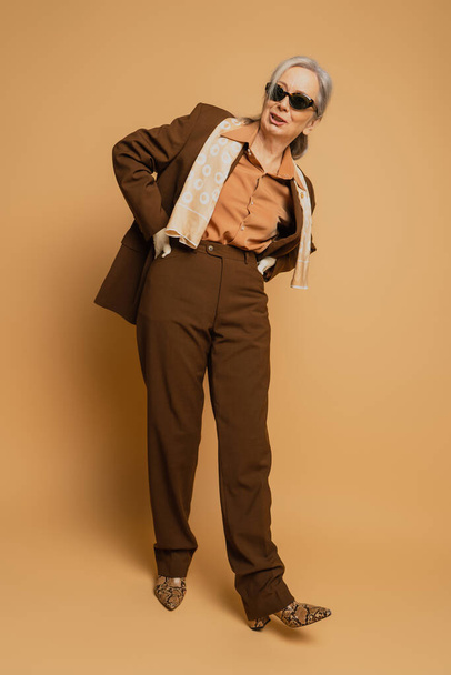 longitud completa de la mujer mayor con estilo en gafas de sol y ropa formal posando con las manos en las caderas en beige  - Foto, imagen