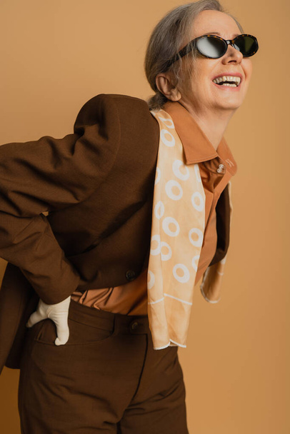 χαρούμενη και ηλικιωμένη γυναίκα με γυαλιά ηλίου και κοστούμι που ποζάρει με το χέρι στο ισχίο απομονωμένη σε μπεζ  - Φωτογραφία, εικόνα