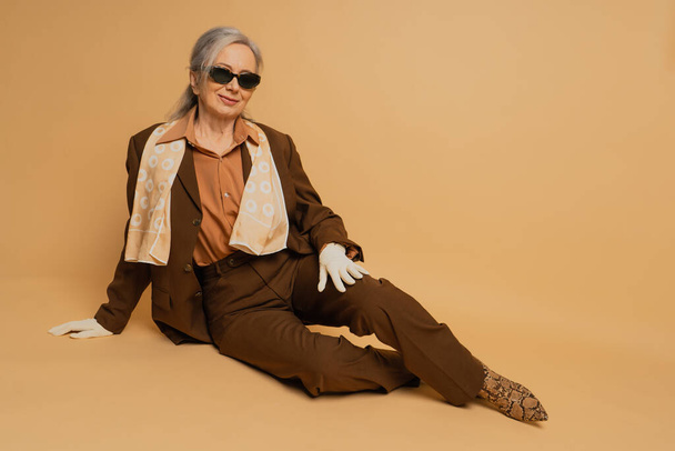 полная длина улыбающейся пожилой женщины в солнечных очках и костюме сидя на бежевом фоне  - Фото, изображение