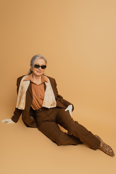 full length of cheerful senior woman in sunglasses and suit sitting on beige background  - Foto, Imagen