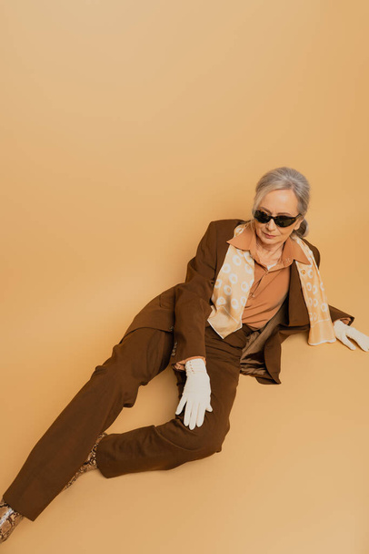 vista ad alto angolo di elegante donna anziana in occhiali da sole e tuta seduta su sfondo beige  - Foto, immagini