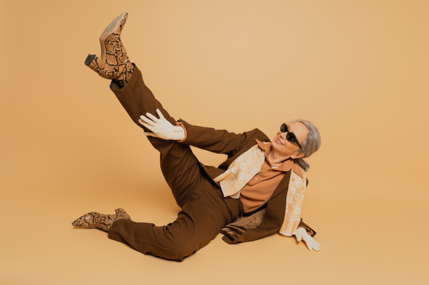 полная длина счастливой пожилой женщины в стильных солнцезащитных очках и костюме позируя с поднятой ногой на бежевом фоне  - Фото, изображение