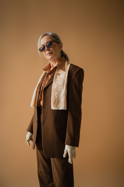 senior woman in brown suit with white gloves and stylish sunglasses posing on beige  - 写真・画像
