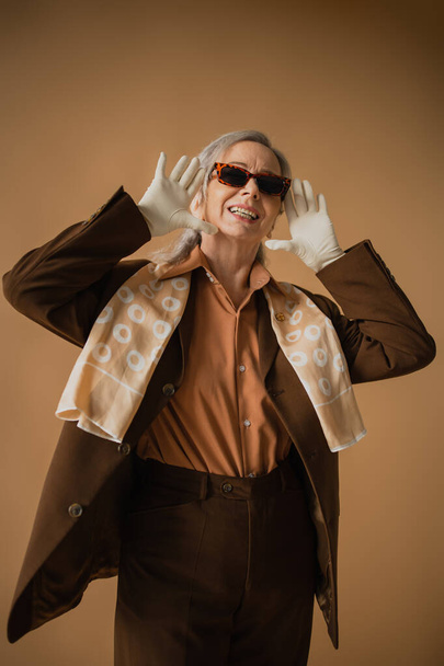 χαμογελαστή ηλικιωμένη γυναίκα με καφέ κοστούμι και λευκά γάντια φορώντας κομψά γυαλιά ηλίου σε μπεζ  - Φωτογραφία, εικόνα