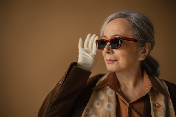senior woman in brown suit and white gloves adjusting trendy sunglasses on beige  - Photo, Image