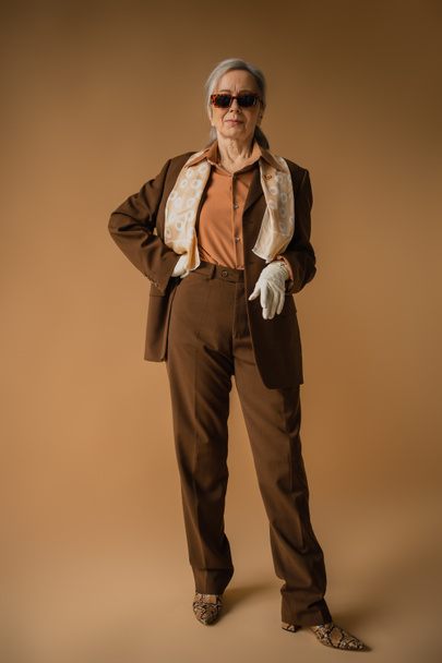 volle Länge der Seniorin in braunem Anzug und weißen Handschuhen posiert mit der Hand an der Hüfte auf beige  - Foto, Bild
