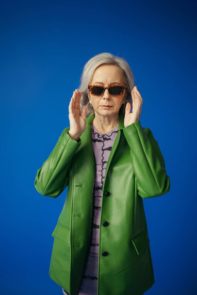 senior woman in green leather jacket adjusting trendy sunglasses isolated on blue - Foto, imagen
