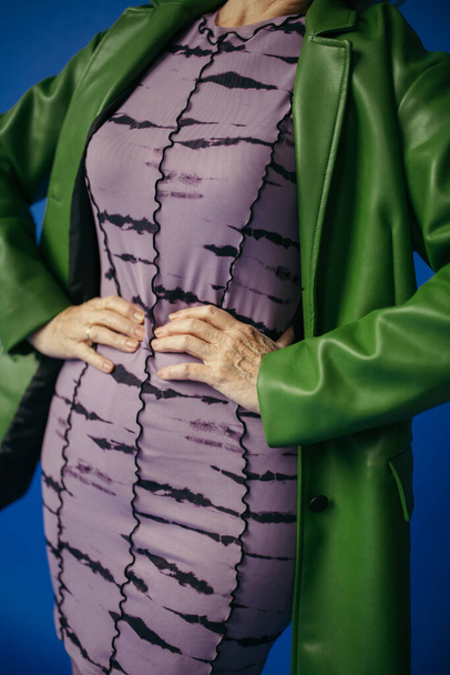 cropped view of senior woman in green leather jacket and dress standing with hands on hips isolated on blue - Photo, Image